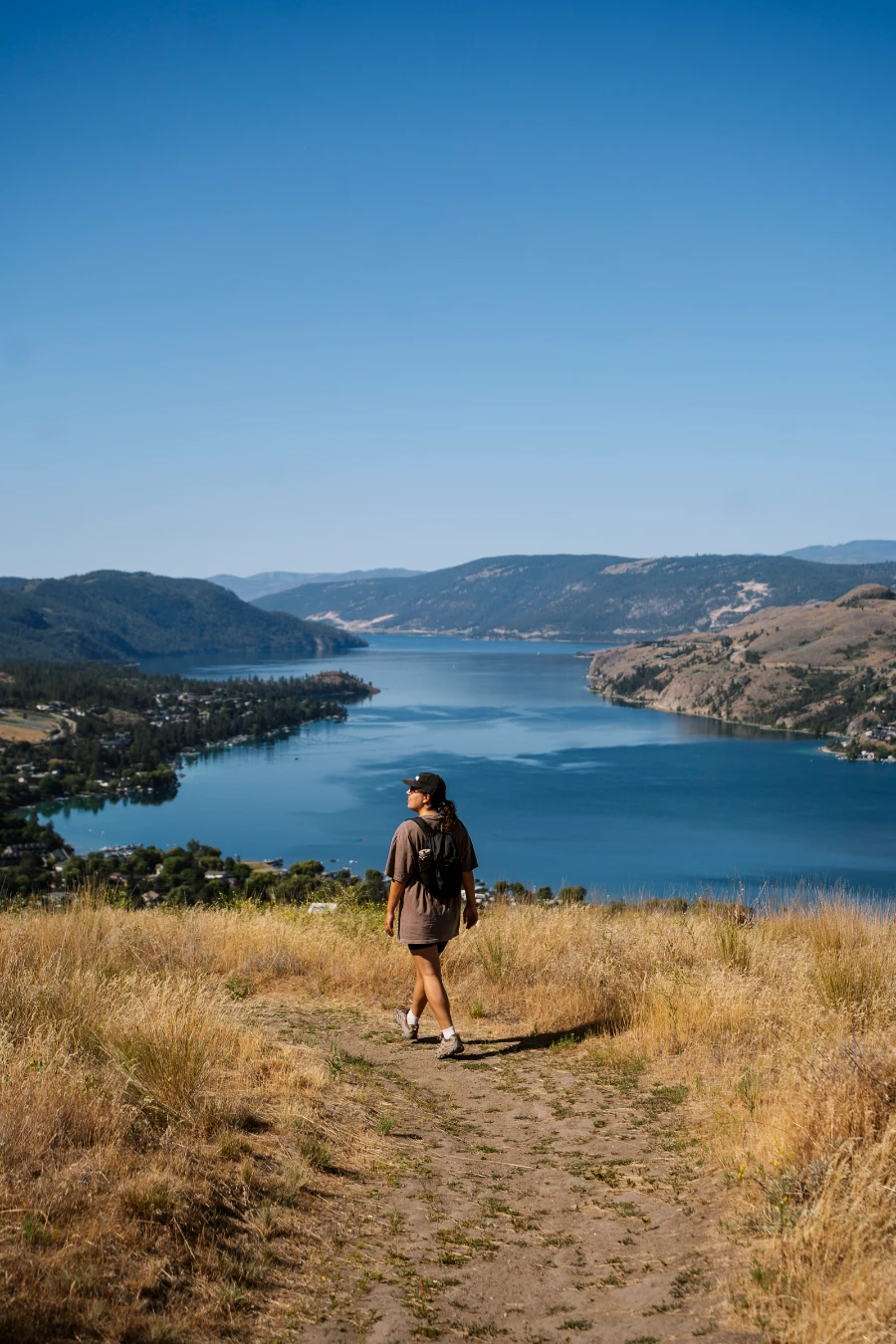 A woman walks along a narrow dirt path with tufts of short grass growing in spots and tall dry grass growing freely on either side, with the large blue mass of Kalamaka Lake in front of her