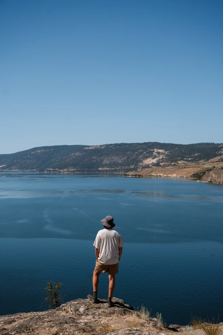 A man stands on a boulder at the highest spot on Rattlesnake Point looking out at the vast blue mass of Kalamaka Lake