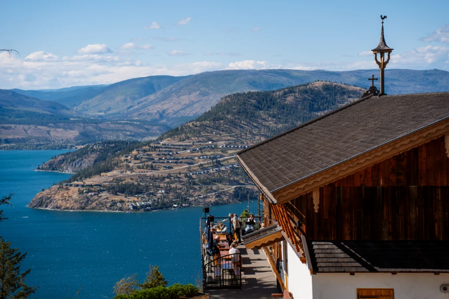 An Austrian-style farmhouse with a wrap-around balcony sits high above the blue waters of Okanagan Lake with rolling hills beyond