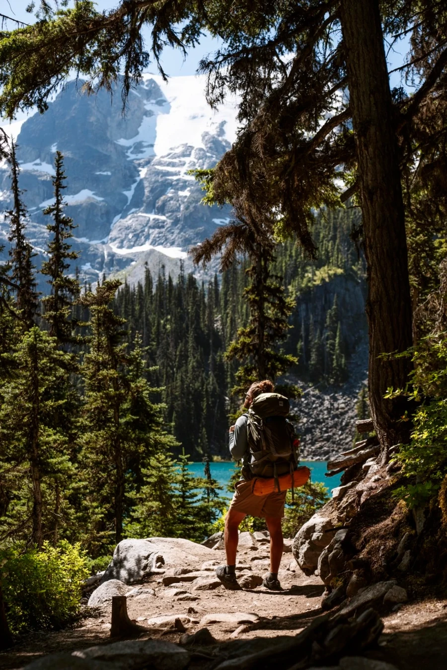 A man carrying a backpacking backpack stands on a stone-covered trail looking out to a blue lake with a snowy mountain towering over the scene