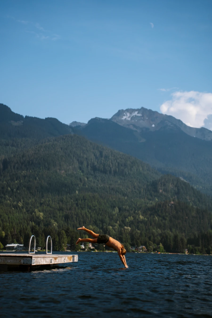 A man diving off of a floating wooden dock with Blackcomb Mountain looming over the lake