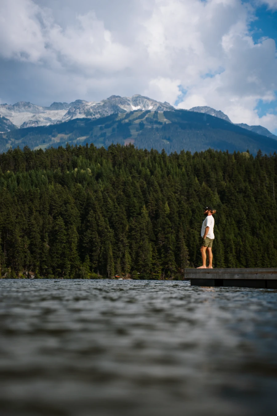 A man standing on a floating wooden dock with Whistler Mountain looming over the lake