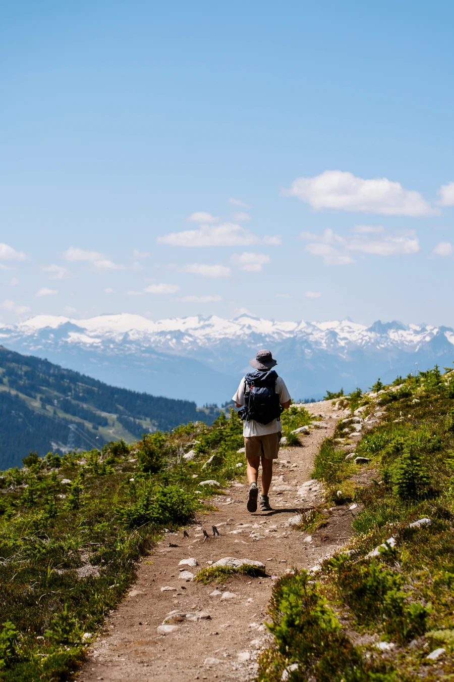 A man with a hiking backpack walks along a wide dirt path with several snow-capped peaks in the distance