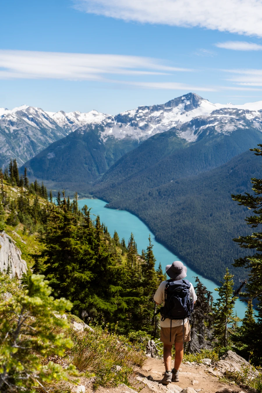 A man with a hiking backpack walking along a narrow dirt trail high above the deep blue water of Cheakamus Lake