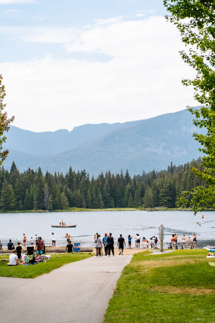 A paved walkway leads past a manicured lawn area towards the sandy beach and wooden dock at Alpha Lake Park in Creekside
