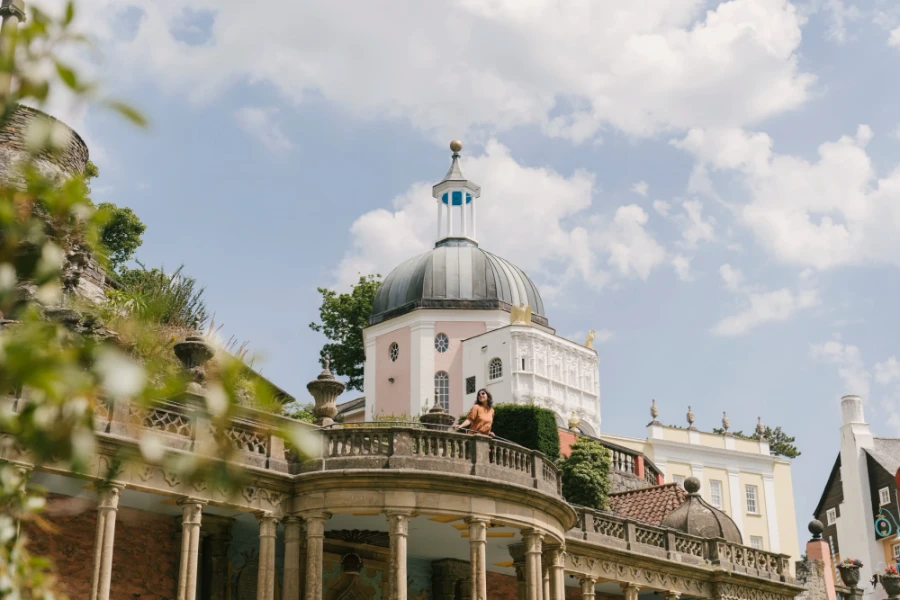A woman stands leans out over a stone veranda with a large dome shaped building behind her in Portmeirion, Wales