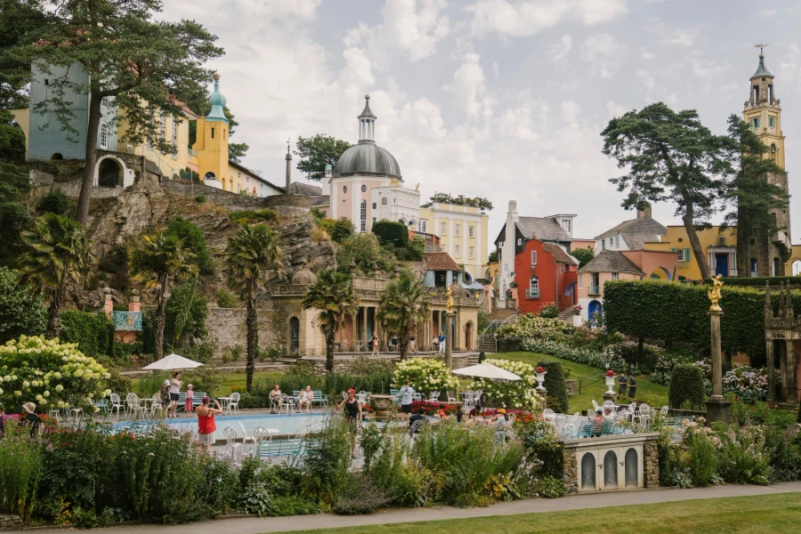 Groups of tourists gather around a garden fountain taking photos with an eclectic collection of buildings built up the hill behind them in Portmeirion, Wales