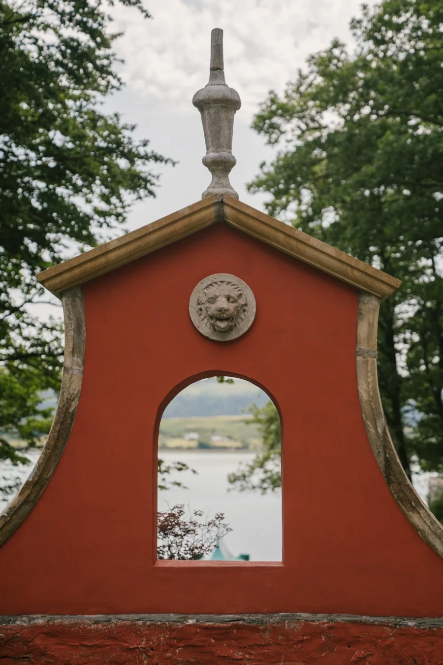 A stone statue of a lions head sits above an archway that looks out to the sea and the farmlands across it in Portmeirion, Wales