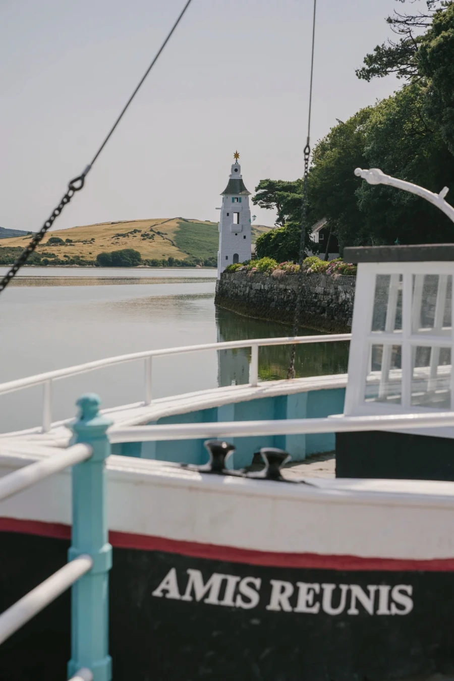 A boat with text on its side reads Amis Reunis and sits docked along a rocky shore with a white lighthouse behind it in Portmeirion, Wales