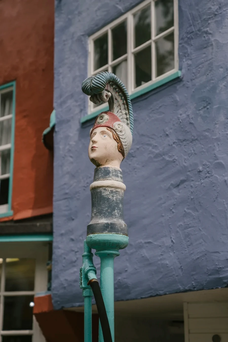 A statue of a womans head wearing a large feathered hat sits atop a water pump in Portmeirion, Wales