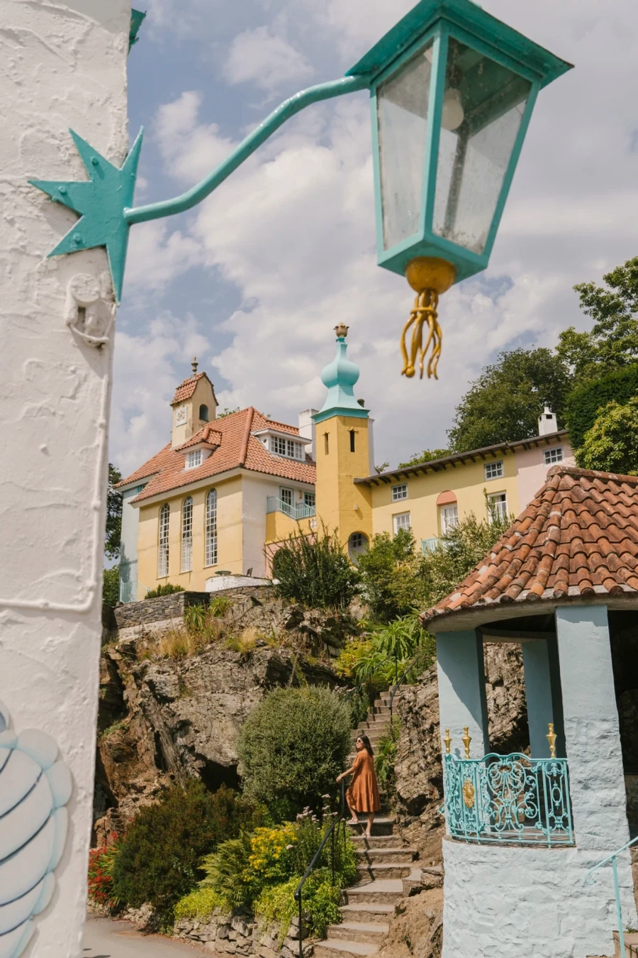 A woman stands on a staircase that leads past a stone tower up to a large pastel colored building in Portmeirion, Wales