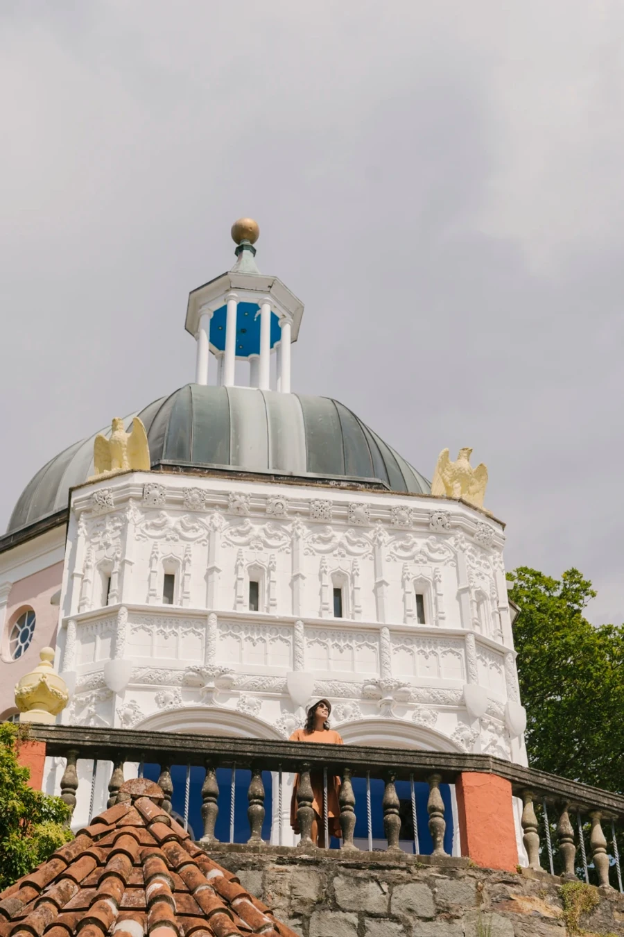 A woman stands above on a stone balcony with a large dome topped building behind her with a detailed ornate white facade