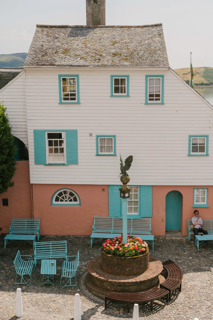 Baby blue benches scattered around a circular flowerbed that has a pillar rising from its centre with a statue of a bird on top in Portmeirion, Wales