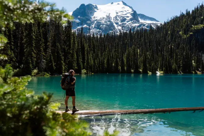 A woman with a large backpack standing on a large log that is floating in Middle Joffre Lake
