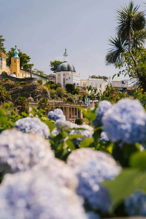 A woman in an orange dress walks down a paved flower-lined walkway in Portmeirion, Wales