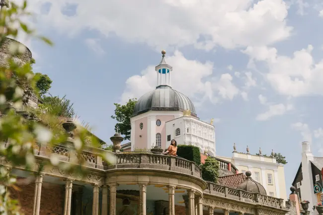 A woman stands leans out over a stone veranda with a large dome shaped building behind her in Portmeirion, Wales