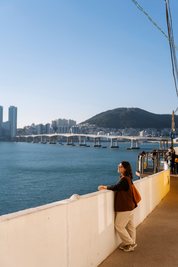 Woman leans on a stone wall above the water with the Busan skyline on the horizon