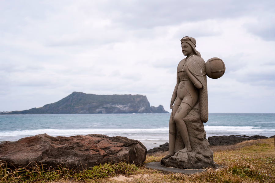 A stone  Hanyeo Statue On Jeju Island sits in front of the sea with another island in the distance