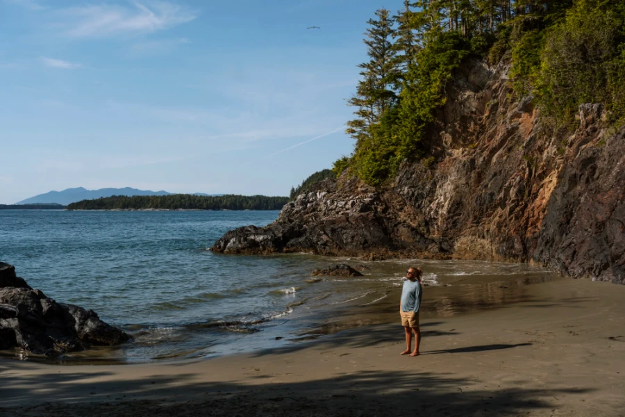 Man standing on a sandy beach with a large stone cliff behind him along the Tonquin Trail in Tofino