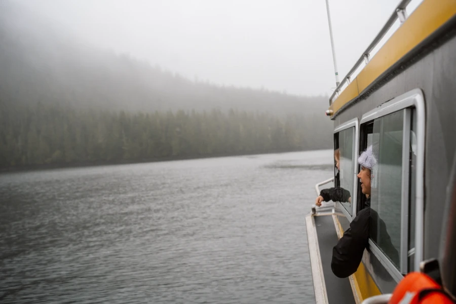 A woman puts her head out of the window of a covered boat to look out across the water to the misty tree-lined coast