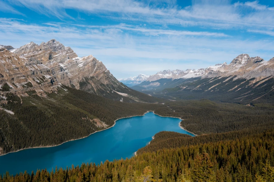 A long narrow blue lake is surrounded on all sides by dense forests that rise up the slopes of the mountainside, above which sit sharp stony peaks