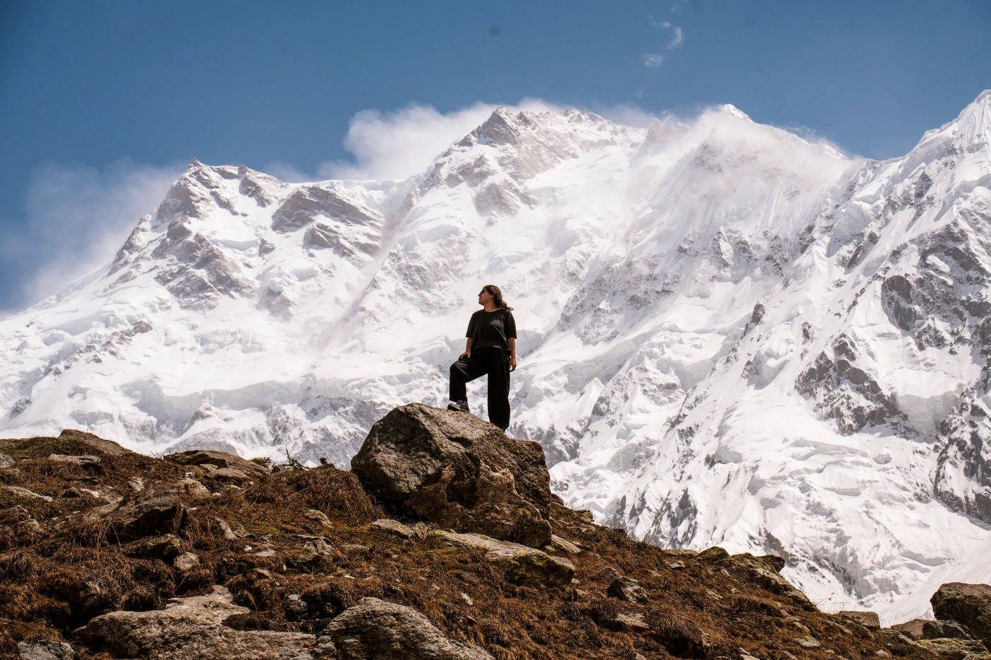 A woman stands on a boulder on a ridge with a massive snow-covered mountain range rising in the distance behind her