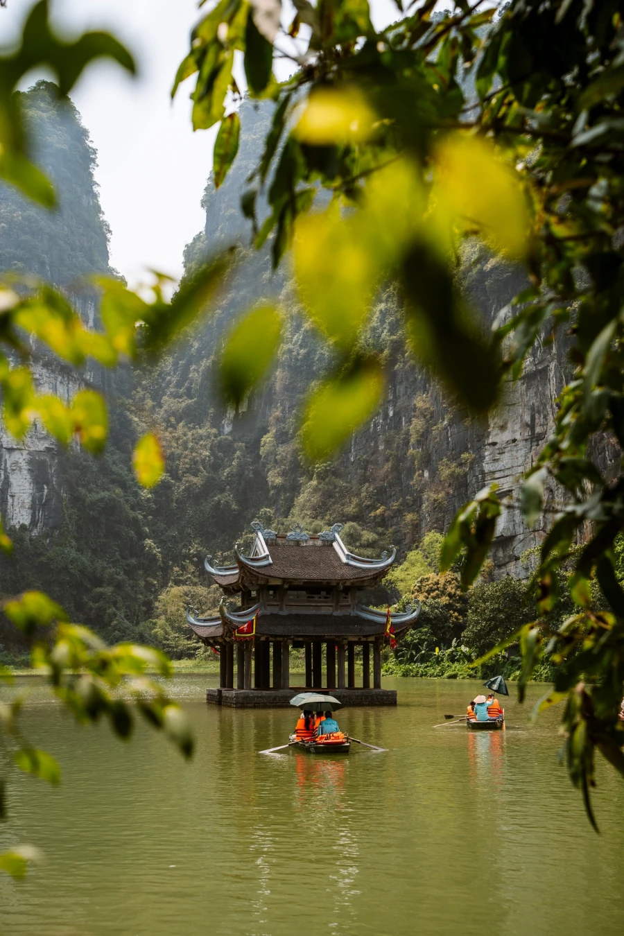 Hanging leaves frame a scene of a calm lake with several boats rowing between the steep jungle mountains and a small temple floating in the water