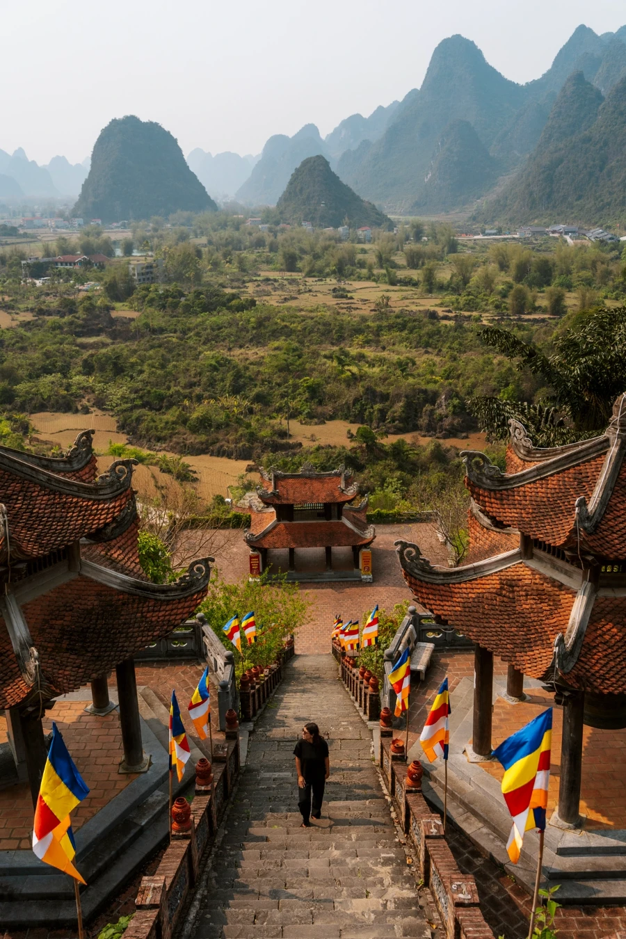 A woman stands on the steep steps between two traditional Vietnamese temple buildings high above a lush valley where several conical mountains rise from the earth