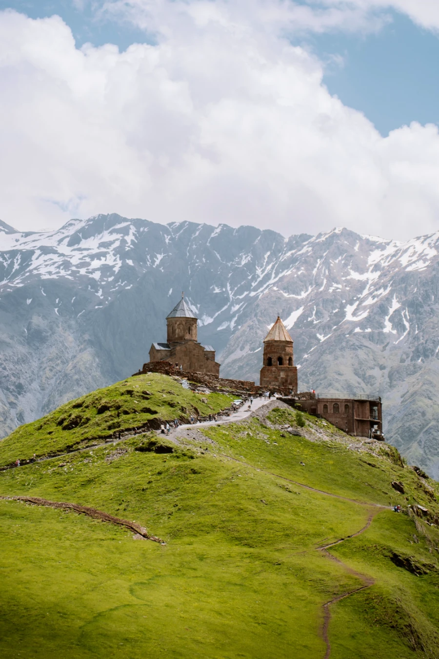 A wide mountain ridge covered in short grass leads to an old-looking castle with two domed towers, with larger snow-capped mountains looming behind
