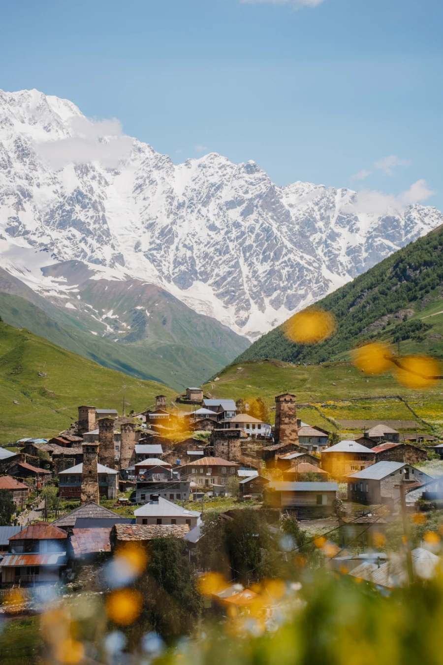 An old stone village nestled into a lush green valley with green mountains rising on either side and massive white snow-covered ridges looming in the distance