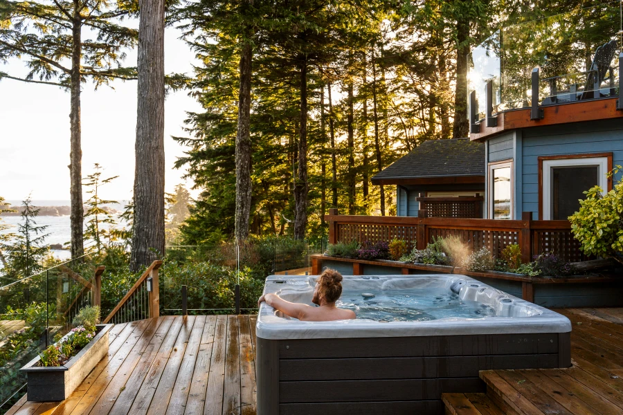 Man sits in a hot tub on a wooden deck surrounded by trees looking out to the ocean in Ucluelet, BC