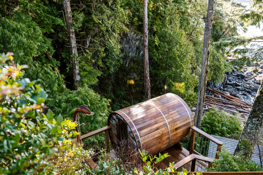A wooded barrel sauna at the bottom of a staircase by the sea and surrounded by trees in in Ucluelet, BC