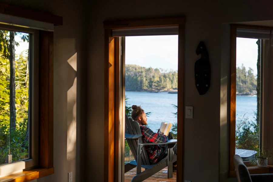 A man reading a book while siting on a wooden Muskoka chair on a deck that looks out to the ocean in Ucluelet, BC
