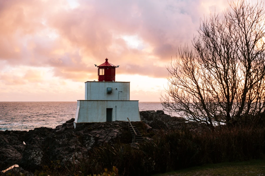 A cloudy sky is illuminated with a pink hue at sunset while a white and red lighthouse sits on a rocky outcrop by the sea c