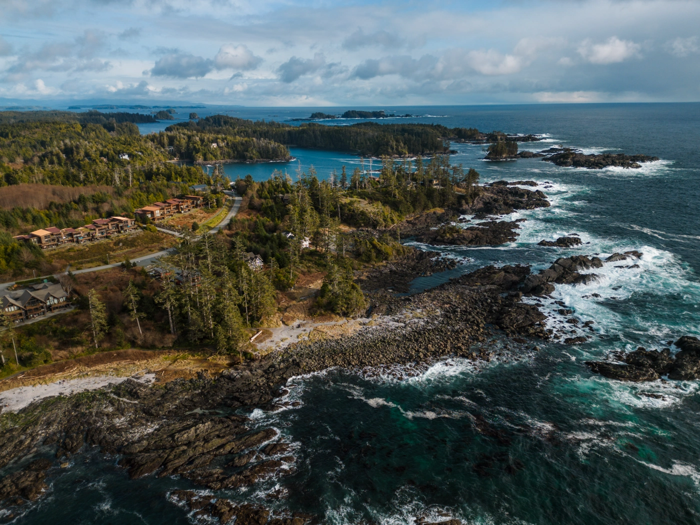 Large waves crash onto the rocky tree-lined shore near Ucluelet with a row of hotels visible beyond the trees