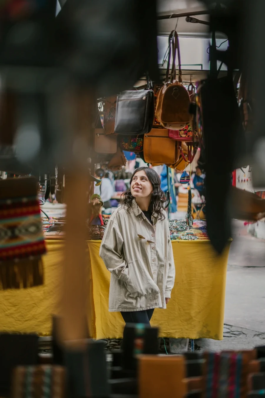 A woman framed by hanging bags and belts looks at tables of goods at Otavalo Market