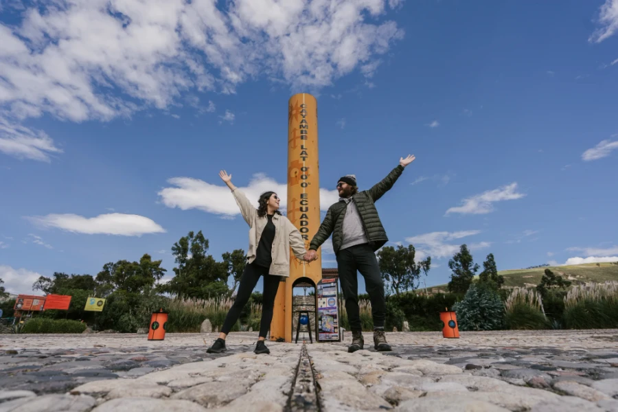 A man and woman stand hand in hand with the woman on one side of the equator and the man on the other with their free arms outstretched