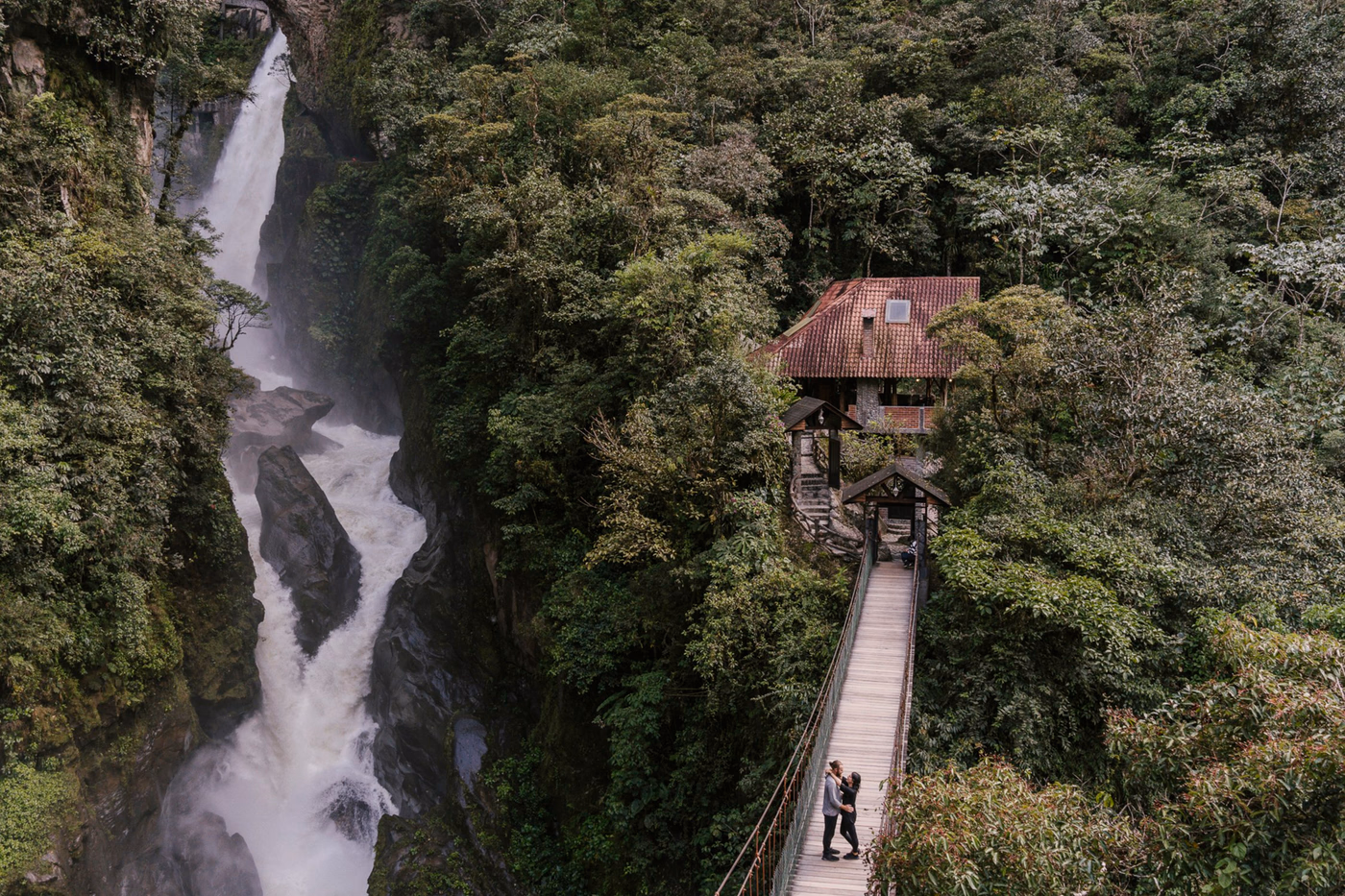 A man and woman embrace in the middle of a suspension bridge with a large powerful waterfall behind them