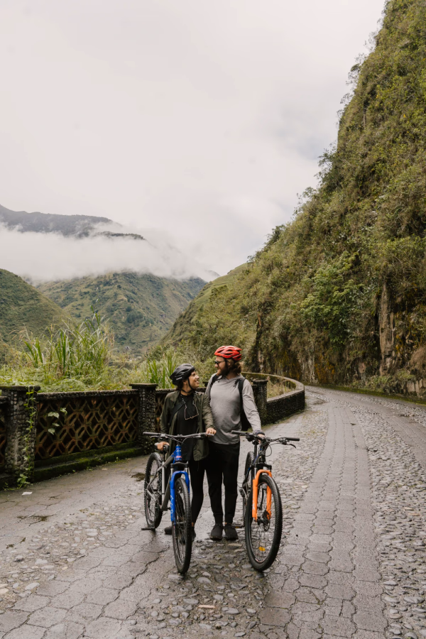 A man and a woman wearing bike helmets stand holding their bikes on a cobble stone road