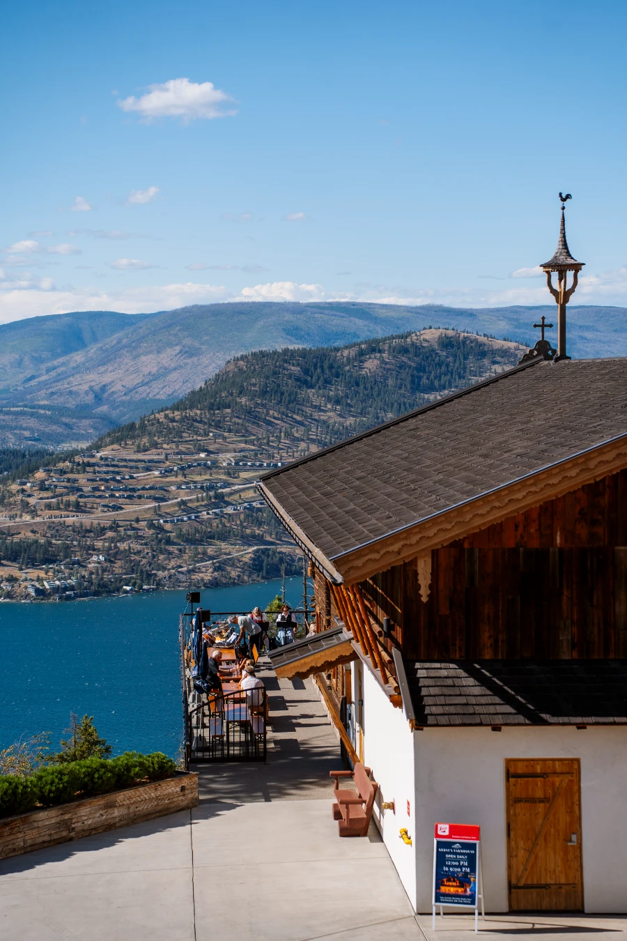 An Austrian-style farmhouse with a wrap-around balcony sits high above the blue waters of Okanagan Lake with rolling hills beyond