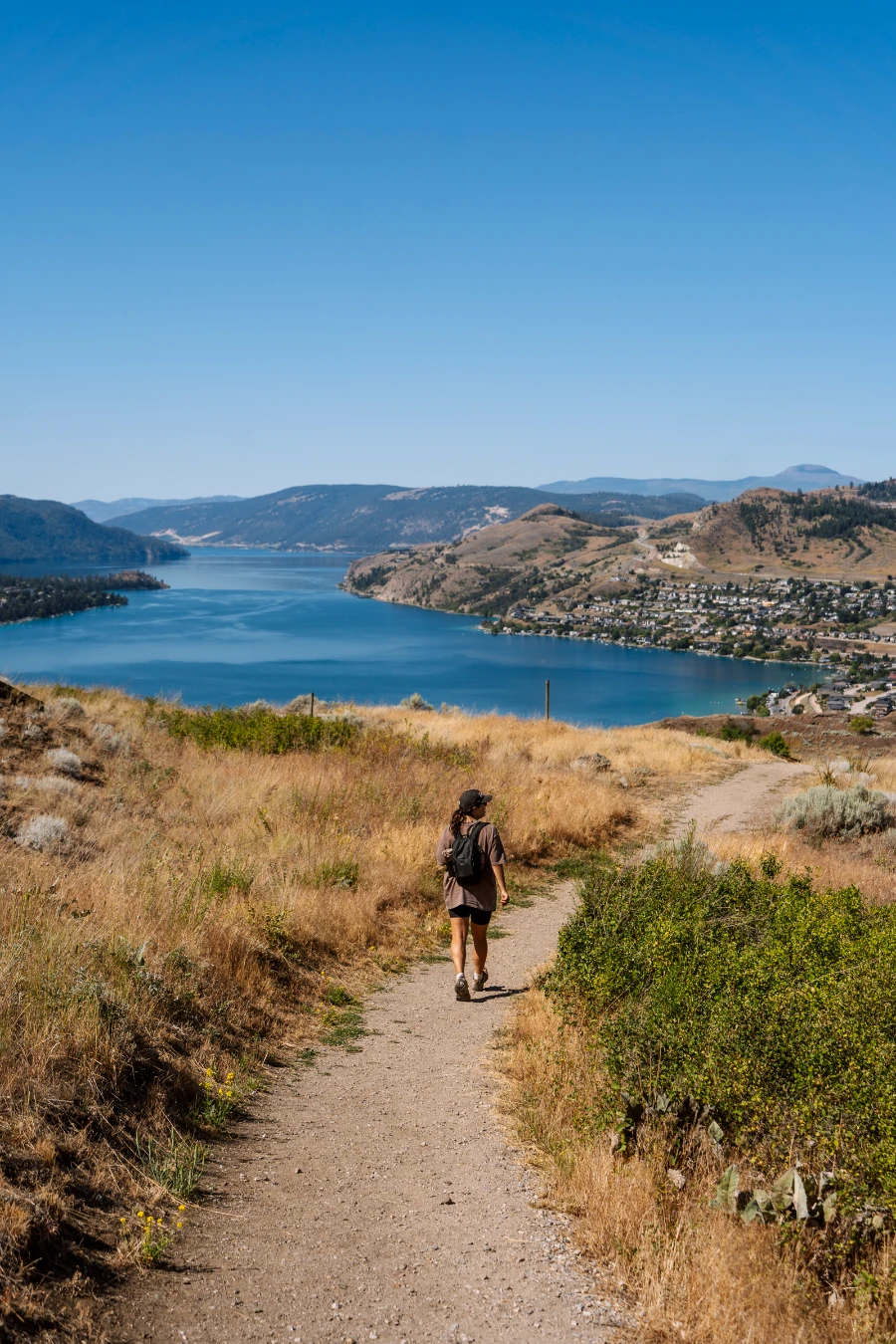 A woman walks along a narrow dirt path with tufts of short grass growing in spots and tall dry grass growing freely on either side, with the large blue mass of Kalamaka Lake in front of her