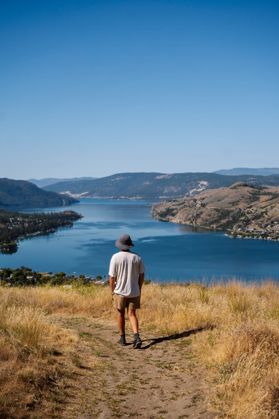 A man walks down a narrow dirt path surrounded on both sides by tall dry grass with the large blue mass of Kalamaka Lake in front of him