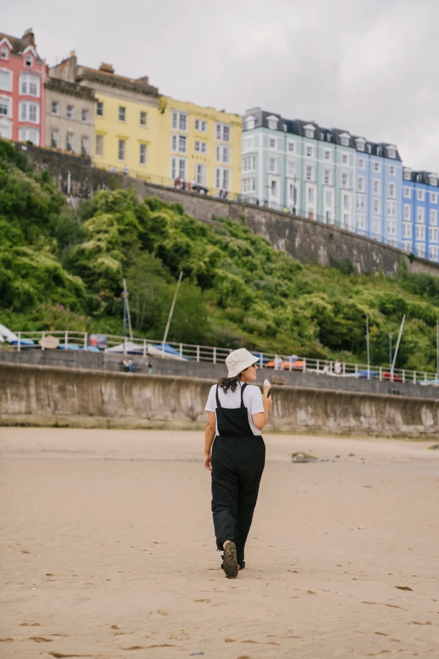 A woman eating an ice cream cone walks along a sandy beach in front a row of multi colored houses