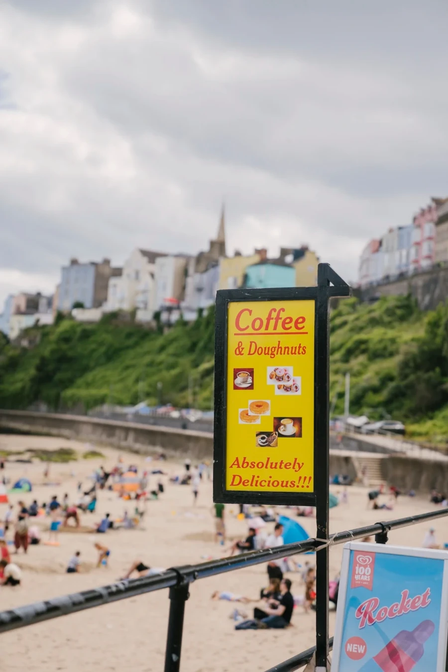 A sign hangs in front of a busy white sand beach reading "Coffee & Doughnuts Absolutely Delicious"