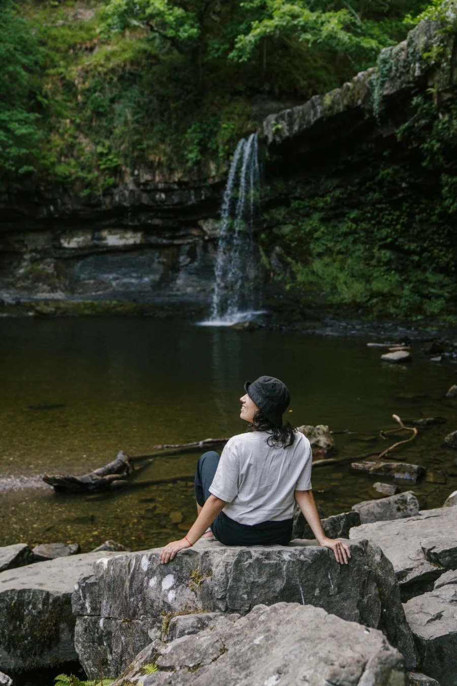 A woman sitting on a rock in front of a pool of water with a waterfall falling into it