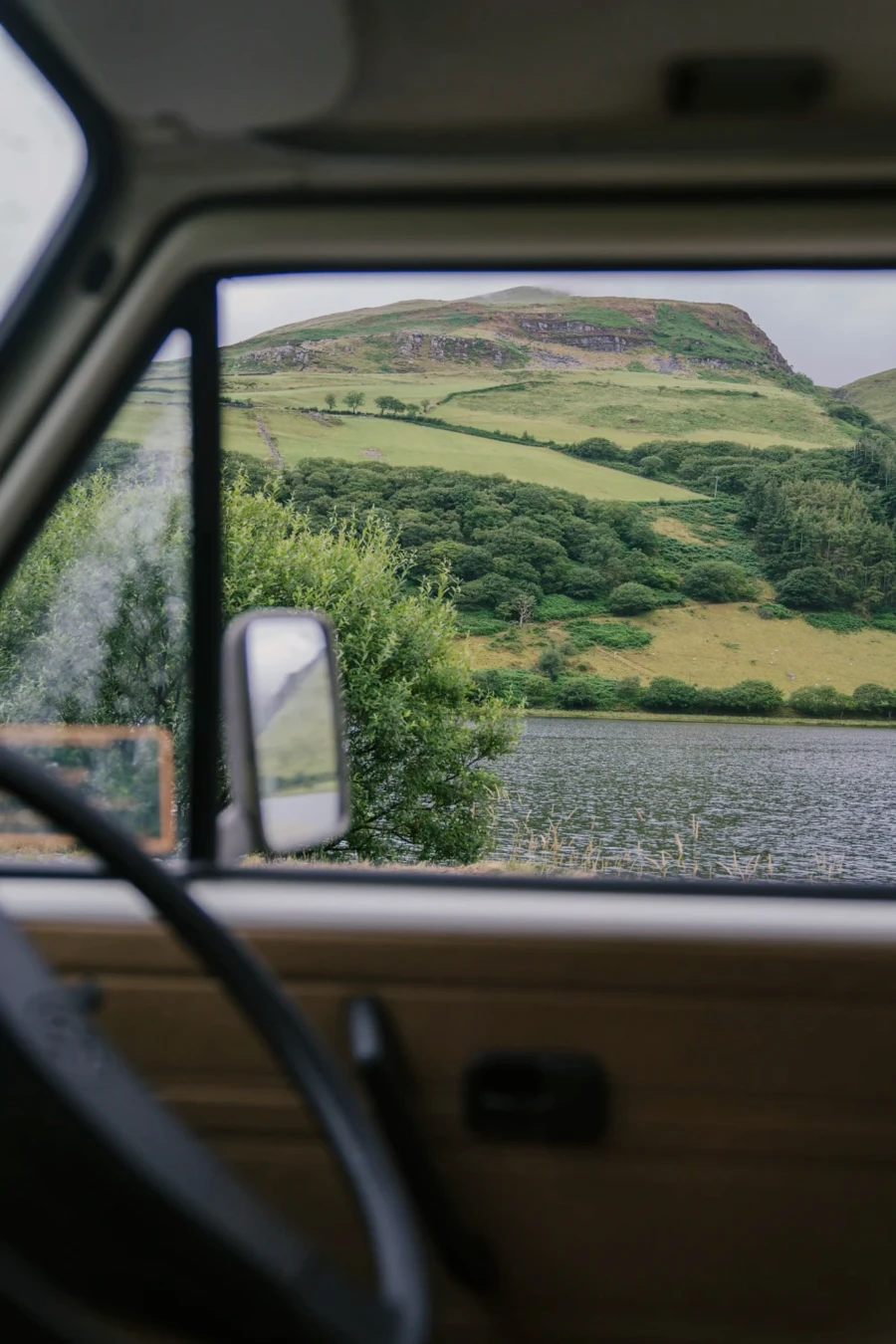 A lake with a forest and mountain behind it framed through the window of a VW Van In Snowdonia National Park, Wales