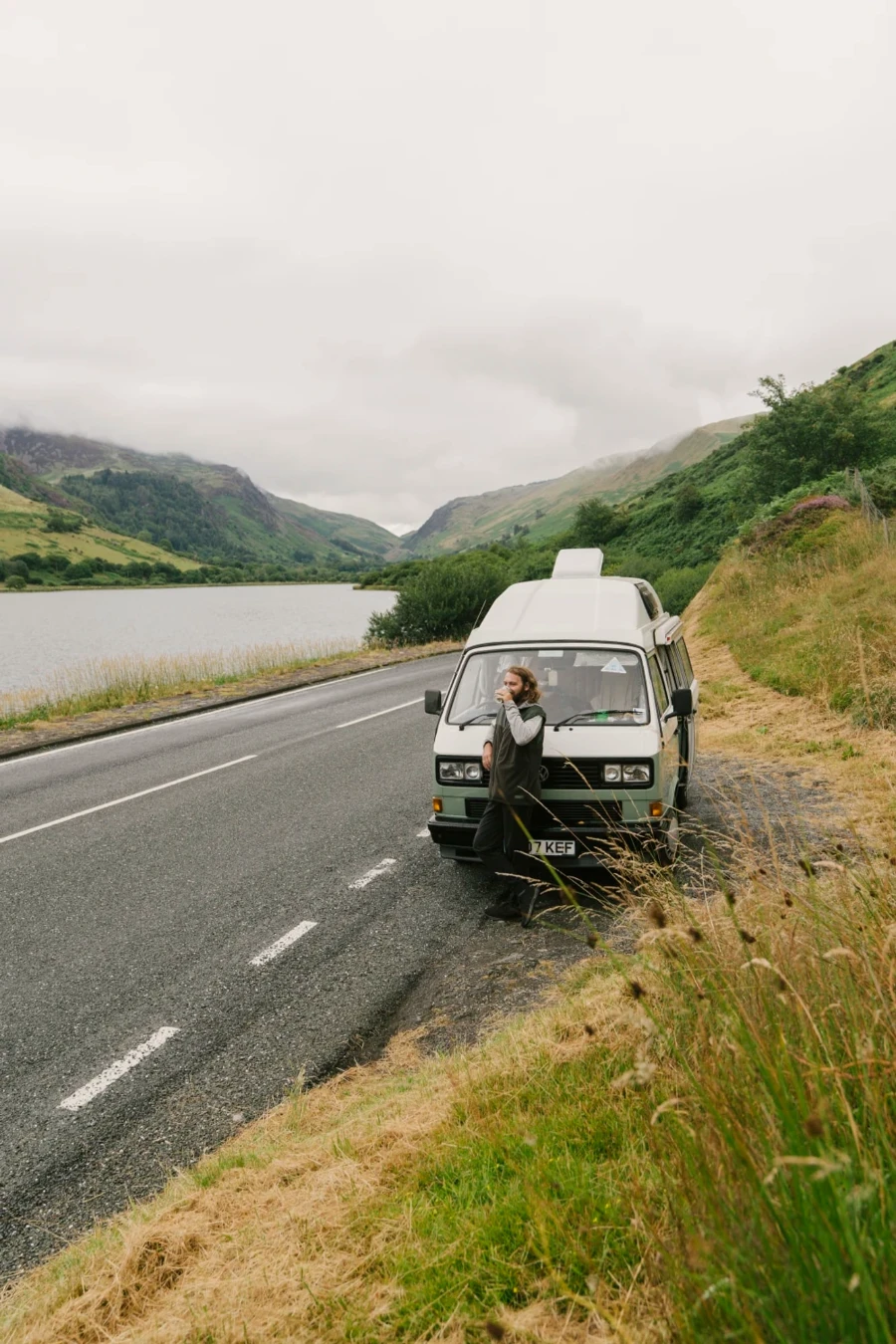 A mean leans against a vw campervan sipping coffee and looking out to a lake in Snowdonia National Park, Wales