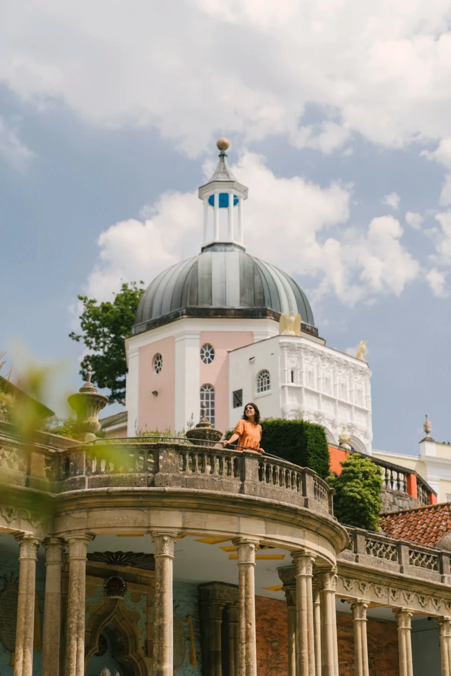 A woman leans on a balcony with pastel coloured buildings behind her in Portmeirion, Wales