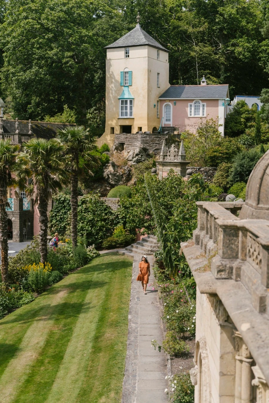 A woman walks along a stone path with manicured grass on one side of here and coloured buildings on the other side