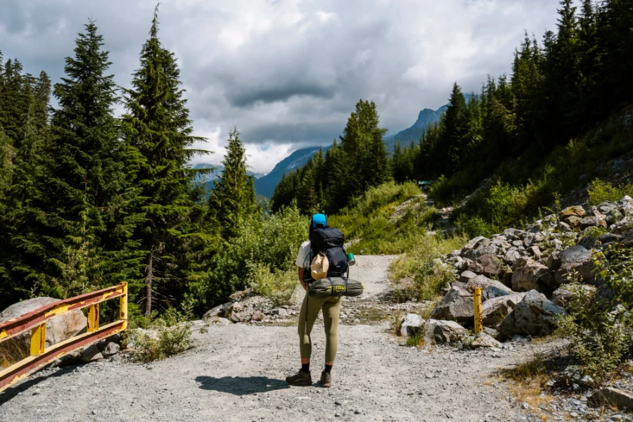 Woman with a large backpack stands on a gravel road near the start of the trail to Watersprite Lake